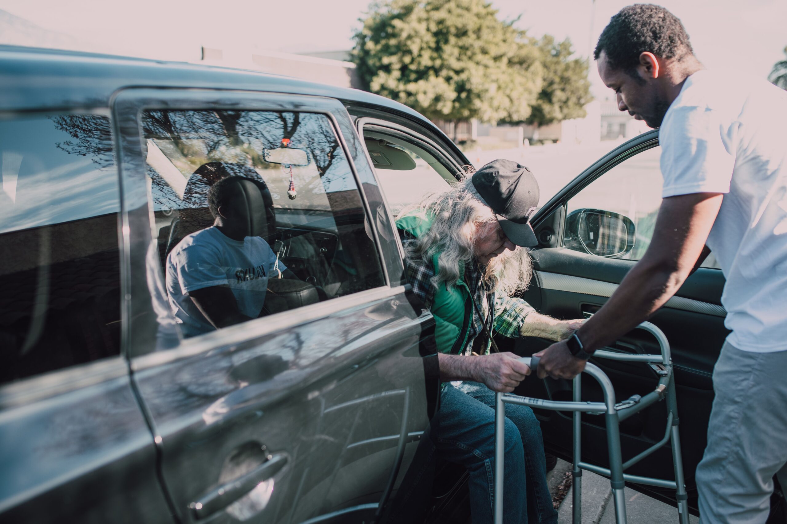 Two men help an elderly person with a walker get out of a car outdoors.